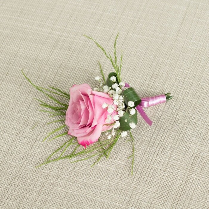 A meticulously crafted blush pink rose boutonniere lies on a pale, textured linen surface, the kind of understated fabric you might find on a carefully set table in a Golders Green family home preparing for a wedding morning. The photograph is taken from above, gently angled so the floral piece appears almost three-dimensional, rising softly from the natural, warm beige backdrop. At its heart is a single rose, partially opened in a perfect balance between bud and full bloom. The innermost petals are a soft, powdery pink, tightly spiralled, while the outer layers loosen into fuller, velvety curves tinged with a slightly deeper, romantic pink along the feathered edges. Surrounding the rose are generous sprigs of baby's breath, each stem dotted with clouds of minuscule, creamy white blossoms that look like a scattering of snowflakes or tiny stars, lending a sense of lightness and air. Fine, grass-like greenery in bright, fresh green fans out from behind the rose, their slim, wispy blades creating a subtle halo and adding movement, as if stirred by a faint breeze drifting down Finchley Road. At the base of the arrangement, a single broader leaf in deep green is folded neatly around the stems, forming a structured cup that gives the boutonniere a polished, tailored finish. The stems are carefully bound with a satin ribbon that runs in two complementary tones: a gentle lavender-pink that echoes the rose's softness and a richer magenta-purple accent that deepens the colour story. The ribbon is tied in a simple, confident knot, without an extravagant bow, reinforcing the design's refined, modern sensibility. Soft, diffuse lighting washes over the scene, creating subtle shadows that accentuate the contours of each petal, leaf, and ribbon fold. Altogether, the boutonniere feels like a small, hand-crafted promise of romance, ideal for a groom or prom guest around Golders Green, lovingly made with the quiet expertise you'd expect from a local florist who understands the area's gentle, understated style.