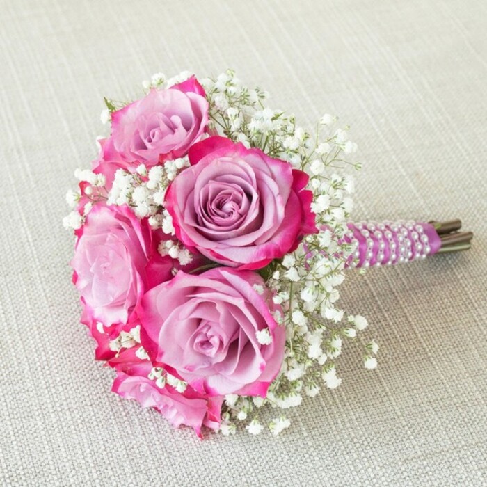 An exquisite five-rose bridal bouquet brought to life by expert florists in Golders Green, this arrangement embodies both modern style and timeless romance. Each focal rose dazzles with a dramatic colour transition-outer petals flush with rich fuchsia pink that melts inward to reveal softened lavender centres, giving every bloom a multi-dimensional depth. Wispy gypsophila, or baby's breath, nestles between the roses, its tiny white florets offering a light, airy contrast that lifts the bouquet and adds visual interest. The compact stems are tightly wrapped in smooth, plum-hued satin, further embellished with several precisely wrapped, spiraling rows of milky white pearls-these details evoke subtle elegance and the bespoke approach typical of local florists near Hampstead Heath. The green stem tips emerge cleanly below the wrapping, signaling both freshness and attention to detail. Set atop a tactile, oatmeal-coloured linen or burlap surface, the bouquet's vibrant tones are beautifully accentuated by the neutral canvas, echoing the tranquil charm of Golders Green's quieter lanes. Perfect for spring or summer weddings, as a bridesmaid's posy or an intimate floral centrepiece, this bouquet reflects the artistry and heartfelt care of Golders Green's floral community, ideal for life's most cherished occasions.