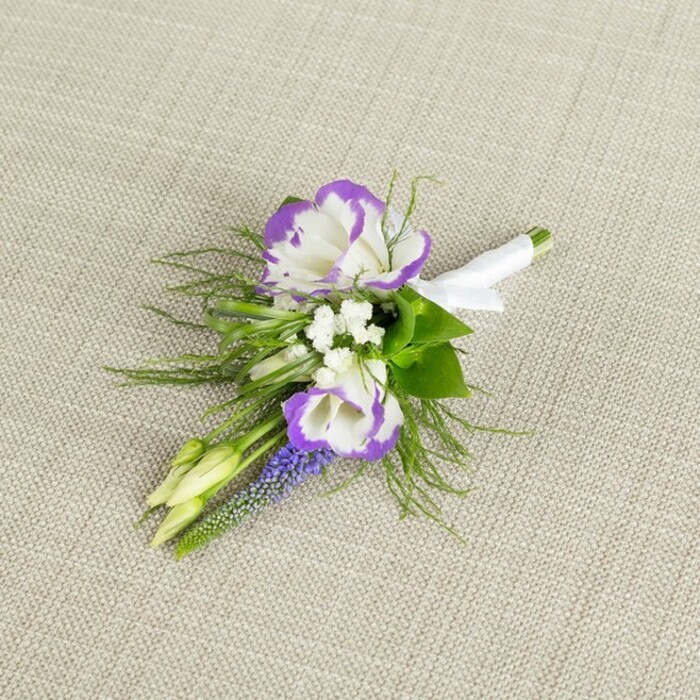 Displayed on a softly textured, light beige linen cloth, this thoughtfully arranged boutonniere feels both classic and contemporary, capturing the sort of understated elegance often favoured at intimate weddings around Golders Green and nearby Hampstead Garden Suburb. At its centre, two lisianthus blooms anchor the design, their petals a pure, fresh white with ruffled edges outlined in a saturated, royal purple that frames each flower like a delicate stroke of paint. These bicolour blossoms offer a striking contrast without ever seeming loud. Around them, tiny white filler flowers, reminiscent of baby's breath, are tucked in gentle clusters, giving a lace-like lightness that softens the stronger hues and creates a sense of air between the main shapes. A slender, upright plume of deep violet muscari introduces a distinctive texture: tightly packed, bead-like bells that draw the eye upward and lend a refined, almost architectural line to the composition. The flowers are embraced by a halo of greenery-a mix of fine, ferny fronds and a few more substantial leaves-which adds depth, movement, and a fresh garden scent you can almost imagine, as if the stems were just conditioned at a local florist's workbench. Two pale green, unopened buds peek from within the foliage, symbolising new beginnings and future moments yet to unfold. The stems are carefully bound and smoothed together at the base with a crisp white satin ribbon, wrapped with the practiced hand of an expert and finished cleanly so it sits neatly on a lapel. The overall impression is one of quiet sophistication, suitable for a groom, groomsman, or prom guest heading to a celebration near Golders Hill Park, looking for a floral detail that complements tailored attire without overpowering it.