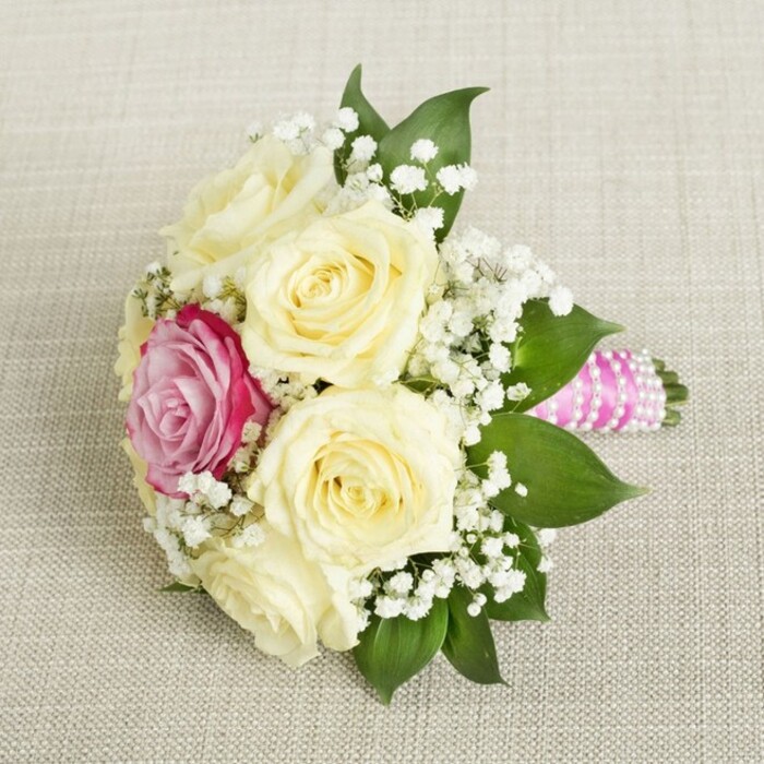 Against a soft, textured canvas reminiscent of the elegant linen used in Golders Green's most charming event spaces, a compact bridal bouquet is artfully displayed from a gently angled overhead view. This arrangement, likely designed for a local bride or bridesmaid, features six pristine cream roses whose sumptuous petals hint at the promise of a fresh beginning. The bouquet's visual centre is a vibrant magenta-pink rose, placed slightly to the left-a bold accent that transforms tradition into a statement of individuality. Wisps of white baby's breath (gypsophila) impart a feathery lightness that dances between the luminous roses, matching the carefree spirit so often found in Golders Green's bustling markets and peaceful garden corners. Framing the florals, thick emerald-green leaves impart depth and balance, offsetting the softness of the petals with a smooth touch. The bouquet's stems are expertly encased in bright fuchsia-pink satin ribbon, wound smoothly to create a polished finish. A string of small, iridescent pearls spirals artfully around the ribbon, sparkling in the light and nodding to timeless bridal traditions. This beautiful, thoughtfully composed piece, resting on a pale linen surface, invites memories of elegant ceremonies and celebratory moments along Finchley Road. Its fragrance-delicate and sweet-combines with tactile pleasures: velvety petals, crisp greenery, and the smooth coolness of pearls and satin. Whether for a wedding at one of Golders Green's local venues or an intimate garden party, this bouquet showcases both professional artistry and heartfelt sentiment, capturing the joy and freshness of life's special occasions.