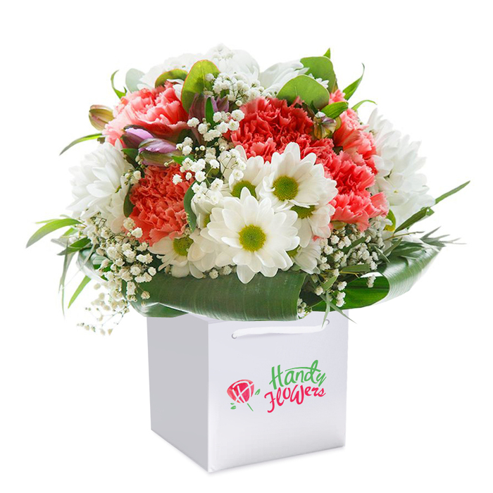 Commanding attention in the centre of the frame, a lavish bouquet nestled within a clean-lined, white gift bag brims with layered colour and organic movement. The arrangement is distinctly abundant, featuring a tapestry of coral-pink and deep red carnations, each blossom identifiable by soft, ruffled petals that impart plushness to the eye. Among these, clusters of bright white daisy-chrysanthemums, their lively lime-green cores glowing, shine as cheerful accents-reminiscent of the local markets bustling nearby Golders Green station. Ethereal white gypsophila weaves delicately throughout the composition, offering an almost magical touch of airiness. Along the left edge, a few tightly closed buds, tipped with purple or mauve, introduce gentle contrast and depth to the otherwise open blooms and recall the intentional detail favoured by Golders Green's community florists. Embracing the bouquet is a collar of broad, gleaming green leaves, framing the arrangement and anchoring it within the square white bag, whose durable white ribbon handles peek from above for practical delivery. The subtly branded bag hints at freshness and elegance but defers to the bouquet's artistry as its focal point. Captured against a pure white background, every flower's hue and texture is celebrated-the textures, the interplay of soft and structured forms, all crafted to suggest a sense of occasion. This ensemble is ready to grace any celebration or special moment, an example of floral design tailored for delivery to Golders Green homes, synagogues, or community gatherings.