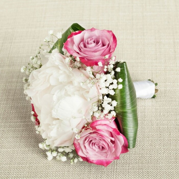 Evoking the quiet elegance of a Golders Green celebration, this sophisticated bouquet features a harmonious blend of classic romance and modern artistry. The composition showcases two captivating pink roses, their petals confidently spiralled from vibrant fuchsia edges to graceful blush-lavender centers, reflecting the unique character of the local neighbourhood. Nestled between them lies a voluminous white peony, each ruffled petal creating a soft, cloud-like texture and a sense of lavish abundance reminiscent of floral displays at Golders Green's local events. Interwoven throughout are delicate sprays of baby's breath, their minute white blossoms bringing an airy, fairy-tale quality that softens the boldness of the main blooms. At the base, folded deep green tropic leaves create an elegant frame, offering both structure and a vibrant contrast, inspired by Golders Hill Park's foliage. The stems are bound with crisp white satin ribbon, forming a neat handle-ideal for a bride or bridesmaid stepping out onto the streets of Golders Green on her special day. Set against a neutral linen backdrop, every petal and detail is softly illuminated, highlighting the freshness and skilled arrangement. This bouquet perfectly encapsulates the blend of refined tradition and fresh creativity that defines Flowers Golders Green, making it a timeless choice for weddings, anniversaries, or as a thoughtful floral gesture.