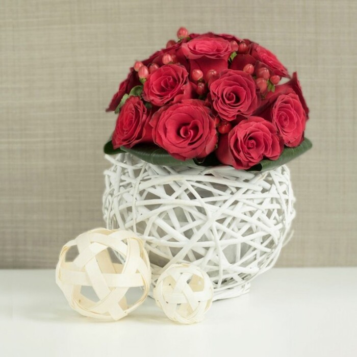 A lavish cluster of rich, crimson roses dominates the composition, carefully shaped into a near-perfect dome that seems to hover just above the table. The roses are tightly packed, their velvety petals overlapping so that only the swirling centres are visible, forming a sea of red that draws the gaze inward. Scattered among the blooms are small groups of red hypericum berries, each berry rounded and polished-looking, providing bright, bead-like accents that play against the lush softness of the petals. At the base of the bouquet, a slim collar of deep green foliage emerges just enough to give a hint of garden freshness, supporting the flowers without distracting from their dramatic colour. This floral canopy is nestled within a distinctive globe-shaped vase, crafted from slender white twigs woven into an open lattice, allowing glimpses of the stems and interior shadows. The white framework feels light and airy, a contemporary touch that would sit beautifully in a modern Golders Green townhouse, perhaps near the leafy calm of Golders Hill Park or the residential streets leading toward Temple Fortune. The vase stands on a smooth, bright white tabletop that reflects a whisper of the arrangement above, adding to the sense of cleanliness and poise. To the left, two decorative balls made from natural, light-toned wicker rest casually on the surface, their tangled strands and circular form echoing the structure of the vase while introducing a softer, more rustic note. Behind everything is a softly blurred, neutral wall in shades of beige and taupe, providing a gentle background that allows the reds and whites to glow. The scene feels tailored for a wedding table arrangement, a romantic dinner at home, or a special celebration with friends gathering in Golders Green after a day in nearby Hampstead Heath.