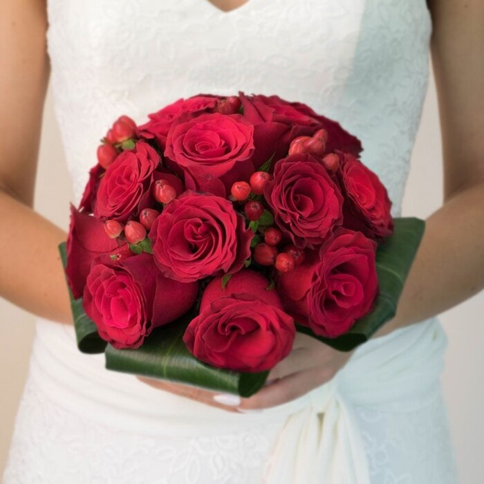 Set against a soft, luminous backdrop mirroring the calm elegance of Golders Green mornings, this close-up, waist-level image draws focus to an exuberant bridal bouquet, lovingly crafted and cradled by a bride in a refined white gown. The wedding dress, adorned with subtle floral lace and a gentle v-neckline, flows gracefully at the skirt, its purity serving as the perfect stage for the bouquet's dramatic palette. Dominating the arrangement, sumptuous deep red roses bloom in harmonious clusters, each flower's densely layered petals imparting texture and visual depth. Nestled skillfully within these roses, shimmering crimson hypericum berries add playful energy and a touch of contemporary flair, their rounded forms like little pearls of happiness. Enveloping the bouquet, gleaming dark green leaves are folded into a meticulously structured collar, reflecting the skill and tradition of local Golders Green florists who treasure their craft. The lighting is gentle, enhancing the tactile richness of the composition, while the bride's neat, manicured hands convey a sense of anticipation and cherished ritual. This bouquet captures a perfect moment of romance and artistry, ideal for weddings across Golders Green, Hampstead, and beyond-where love stories bloom and floral artistry is celebrated.