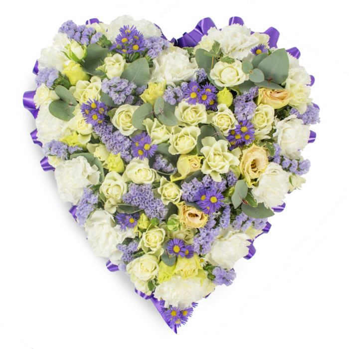 A meticulously arranged heart-shaped floral tribute presented on a plain white surface, the composition feels intimate and reverent: cream and pale yellow roses, many kept as tight buds, form the principal texture, their soft, layered petals creating a luxurious, understated centre. Intermingled are generous clusters of white carnations whose ruffled forms add both volume and a timeless sympathy note, offering a plush backdrop for the subtler colours. Peppering the arrangement are delicate sprays of lavender-hued statice that introduce an ethereal, countryside whisper, while cheerful, small purple asters with sunny yellow centres lift the palette with concentrated accents of richer colour and shape. Smooth, rounded eucalyptus leaves in a dusky blue-green are tucked throughout to create visual breathing room and a tactile contrast; their matte surfaces and sculpted silhouettes calm the overall density. The entire heart is bound and framed by a continuous, looped purple satin ribbon-its deeper tone and lustre outlining the shape with ceremonial grace and drawing the eye around the piece. The controlled soft lighting highlights the tonal gradations from cream through pale yellow to lavender, and the arrangement's compact, skillful construction reads as both artful and respectful. Intended for funerals, graveside remembrances, or memorial services-whether for a chapel near Finchley Road or a quiet family gathering around Golders Green-this tribute speaks of steadfast love, consolation, and the kind of floral care a local florist would choose to honour a life with dignity.