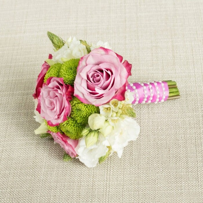 Set against a softly textured, light beige fabric that recalls a natural linen runner laid across a Golders Green dining table, this small bouquet is composed with the precision of a professional wedding design. At its heart, two main roses open generously, their petals silky and layered like folded silk, moving from a gentle pastel pink near the centre to richer fuchsia tones along the ruffled outer edges. A third, slightly more compact rose supports them, giving the composition a sense of depth and progression, as if catching the blooms at different moments in their opening. Encircling the roses are bright lime-green button chrysanthemums, their spherical heads dense with tiny florets that create a distinctive pom-pom texture. These green accents inject energy into the palette, contrasting beautifully with the softness of the pink and the creamy whites scattered through the bouquet. Nestled between the larger flowers are airy sprays of white and cream filler blooms, possibly lisianthus, freesia, and dainty wax flowers, their star-like shapes and feathery buds filling in the spaces without feeling heavy. Subtle touches of darker green foliage provide a natural frame and keeps the arrangement grounded, like a glimpse of gardens off Hamilton Road or around nearby Hampstead. The stems are cut short and gathered tightly, then wrapped in vivid fuchsia satin ribbon that forms a smooth, comfortable handle. Over this ribbon, a fine strand of white faux pearls has been spiralled diagonally, each bead catching the light in a quiet shimmer, adding a bridal jewellery feel to the bouquet. The remaining stem ends are exposed and neatly trimmed. Gentle, even lighting illuminates every petal and texture, suggesting a carefully styled photograph for wedding inspiration or local flower delivery, ideal for a bridesmaid's bouquet, petite bridal posy, or an elegant centrepiece at an intimate Golders Green celebration.