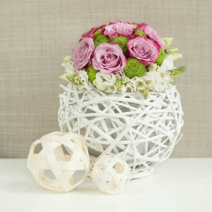 A refined floral display sits on a flawless white surface, reminiscent of a beautifully dressed wedding table in one of Golders Green's boutique venues. At the heart of the image is a spherical white vase, created from intricately plaited branches or rattan-like twigs that loop and cross to form an open framework, giving the container a light, basket-like character while still reading as distinctly modern. Cradled inside this airy vessel is a compact, domed bouquet bursting with fresh flowers. Dominating the arrangement are generous pink and lavender-hued roses, some a gentle pastel blush, others leaning into richer rosy tones, their layered petals curled in perfect spirals that invite the eye inward. Between these larger heads, bright green button chrysanthemums appear like tiny pom-poms, their dense, rounded forms adding both contrast and playful structure. Near the base and occasionally peeking through the roses, smaller white and creamy filler blooms-perhaps spray roses or delicate freesia-introduce a lace-like softness that lightens the overall silhouette and suggests gentle movement. To the lower left of the vase, also resting on the white tabletop, are two decorative spheres crafted from slender strands of natural-toned wicker, one medium and one small, their crisscrossing strips forming open orbs that mirror the texture of the vase without repeating its colour. The background is a warm, muted beige fabric with a subtle woven texture, softly blurred to keep the focus on the flowers and decorative elements. The lighting is calm and even, catching the edges of the petals and the fine lines of the wicker, resulting in an image that feels serene, elegant, and perfectly suited to illustrate wedding centrepieces or refined table styling in and around Golders Green.