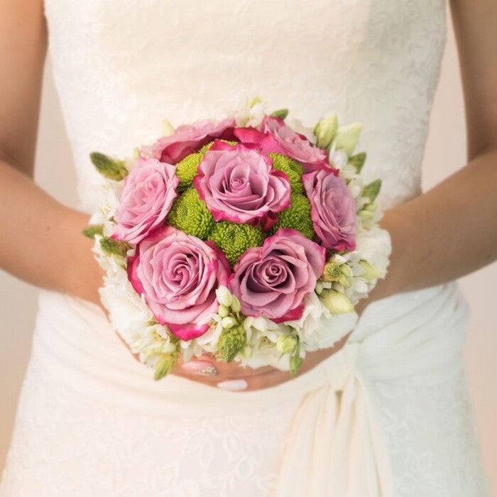 A refined scene in Golders Green unfolds as a bride, poised and serene, presents a compact, perfectly rounded bouquet that radiates sophistication and gentle color play. This close, waist-level view details her off-white bridal gown-its surface traced with exquisite lacework and a graceful, softly draped fabric swath on the right. Resting against this elegant background, the bouquet emerges as the focal point: lush, fully-open roses transition beautifully from pastel lavender and dusty mauve at their core to a more vivid magenta-fuchsia at the petal tips, their depth and vitality enhancing the sense of promise that lingers in the air on Hoop Lane after a summer rain. Intricately placed among these roses, a burst of lime-green button chrysanthemums anchors the arrangement, their playful, rounded shape and fine, dewy petals infusing the bouquet with a sense of new beginnings-echoes of blossoms along Finchley Road in June. Encasing this lively blend, a delicate ring of tiny white flowers-possibly seasonal freesia or compact spray roses-is interspersed with slender green buds and foliage, adding organic energy and soft layers of fragrance. The bride's hands, nails neatly manicured in blush pink with subtle sparkle, suggest both grace and celebration. The gentle light brings out the interplay of textures-lace, petal, bud-turning the arrangement into a story of local craftsmanship and wedding joy, ideally suited for Golders Green couples seeking personal elegance on their wedding day.