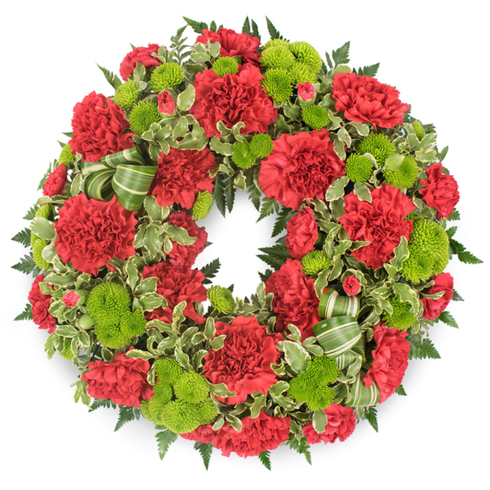 A traditional circular wreath, designed as a funeral or memorial tribute, appears suspended against a bright white background, its colours of deep red and fresh lime green emerging vividly from the frame. The ring is packed from edge to edge with flowers, so no bare space shows through, creating a sense of abundance and care. Dominating the design are red carnations in various stages of bloom, their plush, frilly petals layered densely to form full, rounded heads. The red is a rich, dignified crimson, reminiscent of velvet theatre curtains at an evening service, and each blossom contributes to a soft, gently undulating texture around the circle. Punctuating this sea of red are clusters of lime green button chrysanthemums, each bloom a compact, perfectly shaped globe with minutely detailed florets that lend a smooth, almost moss-like feel. They brighten the composition, giving the wreath a subtle energy that can bring comfort at a graveside in Hoop Lane Cemetery or during a farewell gathering along Finchley Road. Beneath and between the flowers, mixed foliage adds depth and contrast: dark, feathery fern fronds reach outward in delicate arcs, while variegated pittosporum leaves, with their creamy edges and darker centres, lace through the design like soft brushstrokes. A green ribbon, patterned with slightly darker stripes, is woven into the lower right quadrant in loose loops that echo the circular form without distracting from the blossoms. The white background and gentle, even lighting highlight every shape and shadow, emphasising the craftsmanship and balance of the wreath. It feels both solemn and quietly hopeful, a carefully composed expression of sympathy from a local Golders Green florist who understands the importance of honouring memory with grace.