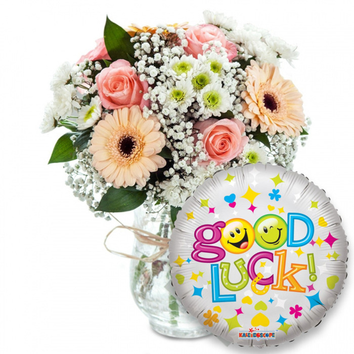 An encouraging 'Good Luck!' gift captured with clarity and warmth: a full, textural bouquet paired with a sparkling celebratory balloon, photographed against a crisp white background so every colour pops. The floral arrangement, set slightly to the left, spills naturally from a clear glass vase tied with rustic twine at the neck; you can see the stems through the glass and imagine the faint, green-smelling oasis within. The flowers are chosen for their comforting tones and mixed textures-soft light-pink roses with velvety petals; cheerful peachy-orange gerbera daisies marked by darker brown centres that give the bouquet visual weight; abundant sprays of frothy white baby's breath (Gypsophila) as delicate filler; and small white and lime-green chrysanthemum florets that add a bright, crisp note. Deep green foliage slips between the blooms, providing contrast and a leafy, slightly resinous fragrance. To the right and overlapping the lower portion of the vase, a round metallic silver foil balloon catches the light, covered in playful stars, hearts and tiny colourful shapes in pink, blue, yellow and orange. The words Good Luck! curve across the balloon in lively, multicoloured letters; the two O's in Good are replaced by jaunty yellow smiley faces-one winking, the other sticking out its tongue-giving the message a warm, human touch. A small KALEIDOSCOPE label is visible along the balloon's lower edge. Whether intended for a student leaving for university, a friend facing interviews, or a neighbour at a Golders Green farewell, this composition feels like a tactile, scented promise of support and optimism.