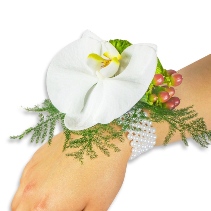 A refined floral corsage displayed on a light-skinned wrist, photographed against a pristine white field so every detail is visible: at its heart, a single, immaculate white Phalaenopsis orchid with broad, gently curving petals and a throat brushed with bright yellow fading into deeper orange, a small sunburst of colour that anchors the composition. Supporting the orchid is a bed of bright asparagus-fern fronds that float outward like soft green lace, their texture providing a natural, cushioning frame. To the upper right, a compact cluster of Hypericum berries introduces a jewel-like sequence of tones-from pale apple green to rose-tinged blush and, at the tips, rich cranberry red-each berry smooth and glossy against the matte greens. Lime-green ruffled accent foliage peeks through, adding crispness and a contemporary edge to the otherwise classic palette. The arrangement is tied to the wrist with multiple strands of luminous white pearls, their tiny spheres catching the light and offering a tactile contrast to petals and foliage; the pearls sit comfortably against the skin, suggesting careful hand-assembly and attention to wearability. The lighting is soft and even, highlighting the orchid's delicate sheen and the fern's filigree without casting harsh shadows. As a local florist's statement piece, it recalls wedding preparations near Golders Green Road and quiet pre-ceremony moments by the cemetery or local synagogue, a versatile boutonnière alternative that balances tradition and modern refinement. Available for collection from Flowers Golders Green, it speaks of fresh materials and careful craftsmanship.