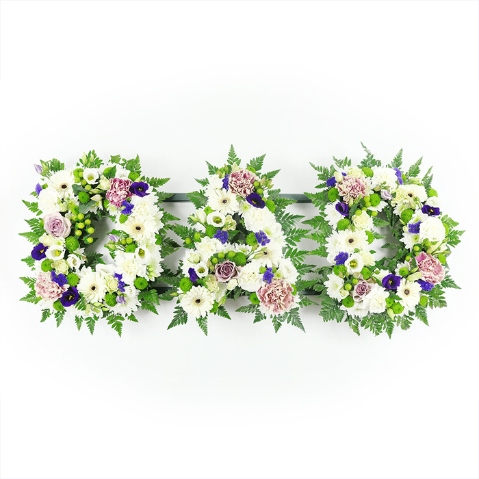 Displayed against a pristine white background, this carefully arranged funeral tribute spells out DAD in three substantial floral letters, aligned side by side in a straight horizontal line. Each letter is densely packed with white flowers, creating a soft yet structured surface that looks almost like a layer of fresh snow, serene and undisturbed. Plump white chrysanthemums form the foundation, joined by neat white gerbera daisies with their distinctive centres and dainty white lisianthus and spray roses that add subtle variation and refinement. The petals, layered and slightly ruffled, give a sense of gentle movement, as though a light breeze might be drifting through them like the air in nearby Golders Hill Park. Around the perimeter and tucked between blooms, lush green fern fronds and other foliage trace the shapes of the letters, the vibrant green echoing the tree-lined roads of Golders Green and adding a sense of enduring life. Interspersed among the white flowers are meaningful touches of colour: clusters of bright green button chrysanthemums bring a lively, fresh contrast, while deeper purples and royal violet tones, perhaps small spray chrysanthemums or pansy-like blossoms, add a dignified richness. Softer lavender and dusky mauve roses and carnations gently thread through the arrangement, introducing warmth and emotional nuance, like shared memories that soften grief. The lighting is bright and even, revealing every tiny detail-from the texture of each petal to the delicate veins in the leaves-without distraction. This DAD floral lettering feels like a bespoke tribute created by a local Golders Green florist, ideal for a funeral cortege travelling along Finchley Road or for placing in a quiet corner of a cemetery, expressing profound love, remembrance, and gratitude for a cherished father.
