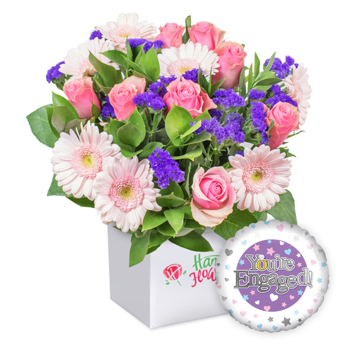 A vibrant engagement bouquet sits in a pristine white square box, the composition showcasing pink roses in full bloom surrounded by light pink and white gerbera daisies whose petals radiate cheer. Quietly dramatic purple filler flowers lend depth and contrast, while abundant green foliage provides a verdant backdrop that makes the colors pop. The container features a subtle logo-a red rose icon with green script-hinting at quality craftsmanship without loud branding. To the lower right, a round foil balloon completes the scene, its central purple circle carrying the words You're Engaged! in elegant silver lettering, accented by a golden O in You're. The outer white ring of the balloon is decorated with a scattering of pastel hearts in pink, blue, and purple, interspersed with tiny silver stars, signaling celebration and romance. The overall image conveys joy and a warm, heartfelt congratulations for a newly engaged couple. In Golders Green and surrounding areas, this thoughtful bouquet is available for dependable same-day delivery to Cricklewood, Temple Fortune, or Hendon, making it easy to share happiness at short notice.