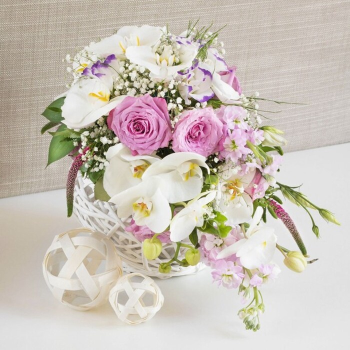 On a clean white surface, a round white wicker basket overflows with a meticulously arranged blend of pink and white flowers, creating a romantic centerpiece that feels perfectly at home in a stylish Golders Green dining room. Dominating the arrangement are generous pastel pink roses, each one fully open to reveal its classic spiral of petals, their soft, velvety texture hinting at a gentle, powdery fragrance. Intertwined with them are striking white Phalaenopsis orchids, their wide, satin-like petals and warm yellow-orange centers adding an unmistakably exotic, high-end flourish. Between these focal flowers, delicate white Lisianthus blooms, some edged with a faint wash of lavender-purple, contribute a ruffled, almost whimsical texture that brings depth and softness. Pale pink stock flowers rise and then arc outward, their clustered florets creating a graceful sense of height and natural movement, as if they were swaying slightly in the light coming through a window off Golders Green Road. Fine sprigs of white gypsophila are scattered throughout like tiny stars, filling any gaps and giving the bouquet a light, airy halo. Lush green foliage peeks from beneath the blossoms and extends in slender trails from the sides, grounding the design with a natural woodland freshness. Beside the basket, on the same white surface, two decorative woven spheres made of pale rattan or wicker add a sculptural contrast, echoing the basket's weave and reinforcing the organic theme. The backdrop is a soft, beige-taupe fabric with gentle texture, reminiscent of a linen curtain, providing a warm, unobtrusive setting that lets the flowers shine. The entire composition radiates refined celebration, ideal as a wedding table arrangement, anniversary centerpiece, or thoughtful gift for same-day delivery around Golders Green and nearby Hampstead Garden Suburb.