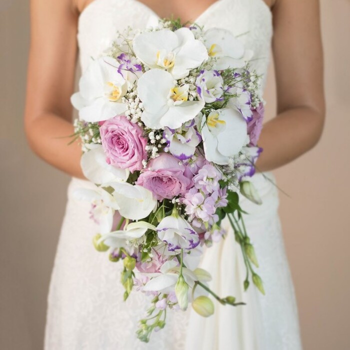 A sumptuous close-up captures a Golders Green bride from the chest down, standing in soft natural light that turns the neutral background a gentle beige, making her opulent bouquet the true focal point. The cascading arrangements flows in a long, elegant teardrop, anchored by creamy white phalaenopsis orchids whose wide, satiny petals and sun-yellow centers speak of purity and quiet grandeur-inviting associations with the serene beauty of Golders Hill Park. Surrounding these regal orchids, clusters of lush, pink garden roses radiate romantic charm, their velvety petals unfurling in intricate spirals. Interlaced throughout the bouquet, lavender stock flowers and delicate purple lisianthus, some tinged with moody amethyst at the edges, lend intriguing pops of colour and soft spiky texture, reminiscent of the ever-changing floral displays on Golders Green Road. Sprigs of tiny, snowy gypsophila add a feathery embrace, ensuring each bloom sits immersed in a cloud of delicate beauty. The bride's simple, strapless white lace gown-its finely patterned bodice subtly visible-emphasises the floral masterpiece held so tenderly in her arms. Minimal green foliage snakes gently through the bouquet, giving just enough contrast to keep the eye wandering downward through the elongated silhouette. This photograph richly evokes the local charm and skilled composition that defines weddings in Golders Green-an extraordinary floral arrangement, timeless in appeal and perfect for wedding inspiration or for couples seeking a distinctive, handcrafted bouquet in the heart of North West London.