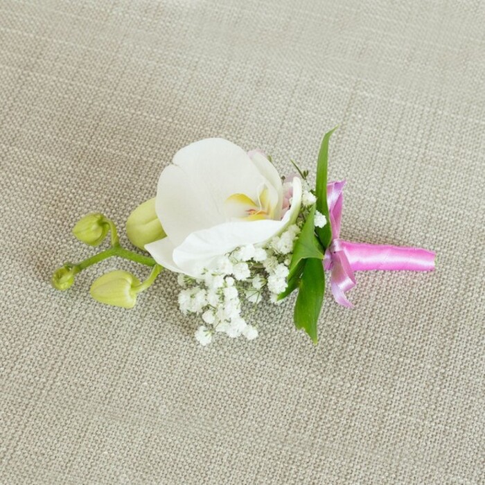 An exquisitely composed orchid boutonniere is shown lying flat on a pale oatmeal, linen-like fabric, the woven threads forming a calm, neutral canvas reminiscent of a finely dressed table in a Golders Green townhouse. At the centre of the composition, a single white Phalaenopsis orchid unfurls its broad, gently arched petals, their surface silky and unblemished. The flower's throat glows subtly with soft yellow tones and the faintest hint of blush pink, drawing the eye inward and adding a delicate warmth to the cool white. Surrounding the base of the orchid, numerous sprigs of baby's breath form a feathery, cloud-soft surround, each tiny white blossom adding texture and volume while maintaining a sense of airiness. From behind the orchid, slender, vivid green leaves emerge, their clean lines and fresh hue offering a natural contrast to the softness of the petals and the neutral backdrop. To one side, a slim stem carries a small cluster of unopened, lime-green orchid buds; their plump, closed forms convey the quiet promise of blossoms yet to open, introducing a gentle narrative of growth and anticipation. The stems of the entire arrangement are neatly held together with a bright fuchsia satin ribbon, its surface smooth and lustrous. The ribbon is tied into a petite bow with short trailing ends that sit tidily along the fabric, adding a sharp, celebratory burst of colour that feels modern yet refined. Soft, even lighting emphasises every curve of the orchid, the tiny spheres of the Gypsophila, and the subtle weave of the background cloth. Crafted with the attention to detail you would expect from an experienced local florist serving Golders Green and nearby Temple Fortune, this floral accessory suits grooms, groomsmen, and special guests at weddings, proms, or elegant evening events across North West London.