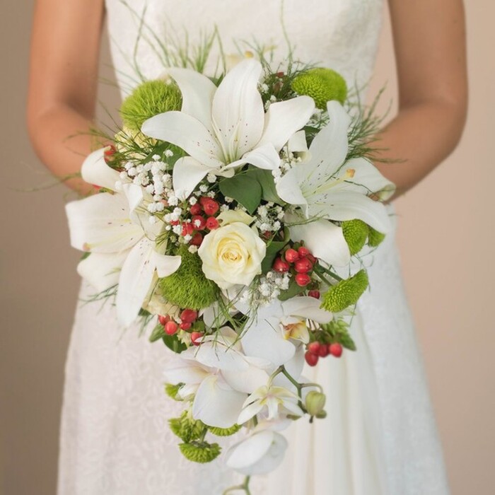 Captured in a sunlit moment in Golders Green, this photo reveals a bride gently embracing a dramatic, artfully cascading wedding bouquet. The arrangement radiates a classic yet contemporary elegance, anchored by several pristine, white lilies with bold trumpet forms and subtle chestnut-speckled interiors. A single, creamy white rose at centre stage evokes the timeless beauty so often celebrated in North West London's leafy avenues. The bouquet's silhouette is elongated and graceful, thanks to the trailing Phalaenopsis orchids whose white petals tumble softly downward, their delicate curves reminiscent of natural water features found in Hampstead Garden Suburb. Interwoven are vibrant lime-green cushion mums, providing a fresh surge of colour and visual texture, offset by cheerful bursts of bright red Hypericum berries reminiscent of the local seasonal markets. Airy clouds of white baby's breath and slender, grass-like streams of foliage enhance the bouquet's structure, giving both movement and a touch of wildness. The visible hint of fine lace at the bride's wedding gown adds an extra layer of intimacy and romance, while the entire bouquet is framed by a background gently out of focus-allowing every floral detail to shine. This luxurious, handcrafted bridal bouquet, inspired by the verdant parks and spirited community of Golders Green, would be a perfect fit for modern weddings seeking both opulence and a nod to classic floral tradition, evoking memories of love and celebration in this distinctive London neighbourhood.