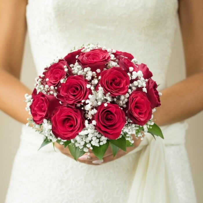 Seen through the refined lens of a Golders Green florist, this tender moment centers on the embrace of a classic, dome-shaped bridal bouquet. The bouquet is a stunning assembly of deep red roses-some buds tightly closed, others unfolding layer by velvety layer-merging gracefully in a cluster that radiates warmth and depth. Scattered amid the blooms, the purest white clouds of gypsophila lend a touch of ethereal lightness and a soft, whispering contrast to the saturated crimsons. Just beneath the florals, a few bright green leaves peek out, subtle reminders of the bouquet's living freshness and the skillful eye that gathered them. The bride's hands, delicately poised with a natural polish, cradle the composition, their reflection of steady anticipation and treasured ritual. Her wedding gown, glimpsed just above the bouquet, features an intricate, likely lace pattern-its detail and subtle shimmer perhaps reminiscent of elegant boutiques lining Golders Green's local high streets. A gentle drape of white fabric falls along her side, echoing the bouquet's sense of graceful flow. The photograph's softly blurred, neutral-hued backdrop ensures that all attention rests on the interplay of rich floral tones and bridal textures. This evocative image captures not only the beauty of expertly crafted wedding flowers but also the community traditions and intimate celebrations that flourish in Golders Green.