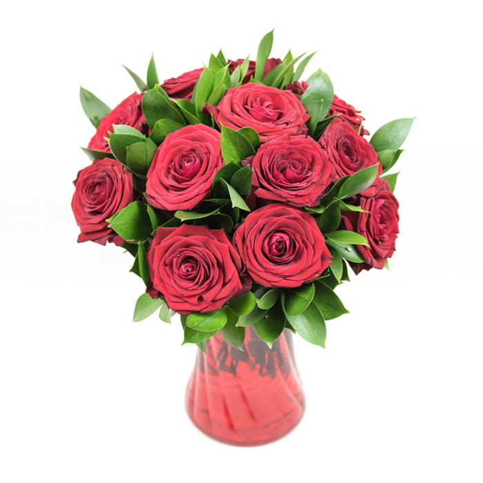 An opulent bouquet of deep crimson red roses is artfully arranged in a translucent red glass vase, standing alone against a pure white background that heightens the drama of its colours. Around twelve to fifteen roses form a dense, rounded dome, each bloom fully open yet still fresh, with thick, velvety petals unfolding in tight, layered spirals. The hue of the petals is a saturated, ruby red, rich and consistent from base to tip, capturing that classic romantic tone often requested by couples in Golders Green marking anniversaries or special evenings. Nestled among the roses is a generous amount of dark green foliage, most likely ruscus or salal, its pointed, glossy leaves threading between the blooms and spilling slightly beyond the circumference of the bouquet. This greenery adds visual depth and variation, the deep emerald tones creating a pleasing contrast that makes the red appear even more intense. The arrangement is supported by a red glass vase with a smooth, gently curving profile that narrows towards the base and widens subtly at the mouth, perfectly proportioned to hold the rounded mass of roses. The glass has a delicate ribbed or swirled texture, catching the bright, even lighting and reflecting highlights so that different shades of red shimmer across its surface. Through the translucent glass, hints of the green rose stems can be seen, grounding the design in natural realism. The crisp white background and balanced illumination mean there are no distractions, only the interplay of red blooms, green foliage, and glass. The composition feels elegant and intentional, a quintessential expression of love, passion, and appreciation, ideal for representing local flower delivery from trusted Golders Green florists to nearby areas such as Temple Fortune and Finchley Road.