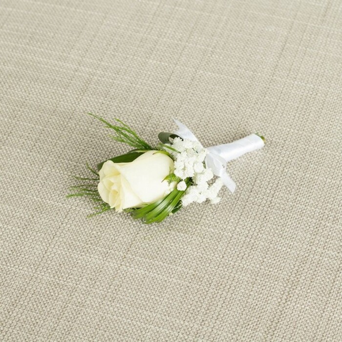 A meticulously crafted white rose boutonniere is displayed at a diagonal angle on a pale beige, linen-like fabric, its fine weave and subtle crosshatch pattern evoking the calm elegance of a thoughtfully dressed wedding suite near Golders Green station. Centered in the composition is a single creamy white rosebud, compact yet full, with petals that are smooth and velvety to the eye, gently layered and still mostly closed, suggesting a bloom at its freshest moment. At the base of the rose, a soft halo of green sepals cradles the flower, hinting at its natural origins. Flanking the rose, dainty heads of white baby's breath cluster together in light, airy sprays, each tiny bloom a pinprick of softness that creates a cloud-like effect around the central flower. Interwoven among the blossoms are narrow, dark green leaves and a few finely textured, fern-like sprigs, which add structure and a rich contrast, reminiscent of foliage found in the shaded gardens and tree-lined streets around Hampstead Garden Suburb. The stems of the rose and greenery are gathered tightly and wrapped with immaculate precision in white satin ribbon, its surface gently lustrous. The ribbon is secured with a small, symmetrical bow placed just beneath the rose head, signifying the piece is complete and ready to be pinned to a groom's or groomsman's lapel. Soft, diffused lighting washes over the scene, highlighting every curve of the petals, the tiny buds of gypsophila, and the satin's gentle sheen while keeping shadows minimal. The boutonniere exudes a mood of timeless romance, purity, and quiet confidence, perfectly suited to formal occasions, registry office ceremonies in Golders Green, or intimate celebrations in nearby West Hampstead.