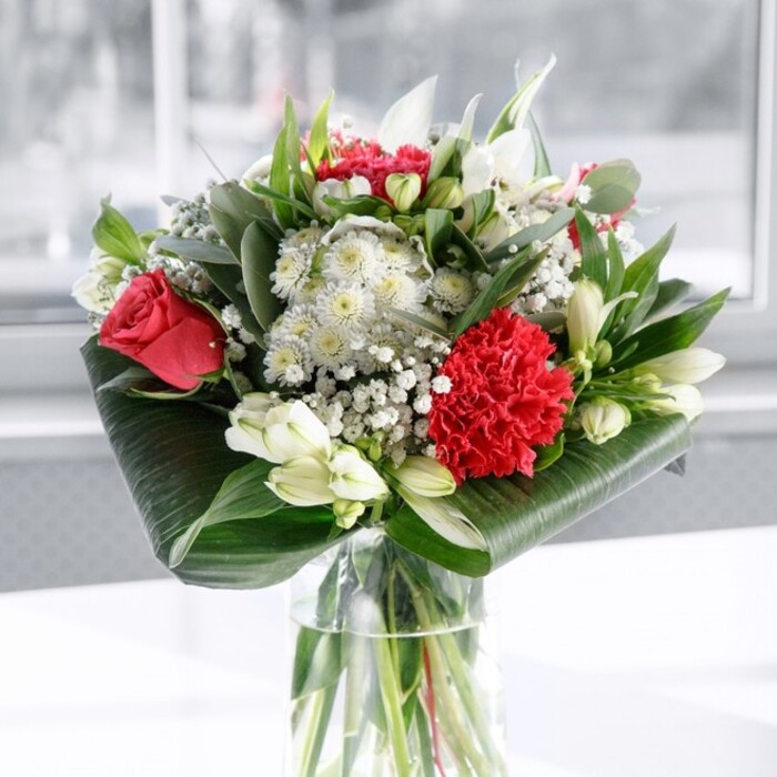 The photograph showcases a luxurious bouquet in a clear cylindrical glass vase, designed in rich red and crisp white tones that echo classic romance with a modern Golders Green twist. At its centre, several deep red roses unfold slowly, their velvety petals layered in perfect spirals that draw your attention inward, like the quiet pull of a familiar Hampstead Garden Suburb walk. A large, deeply ruffled crimson carnation rests near the front, its textured petals creating ripples of shadow and light, adding a sense of depth and movement to the arrangement. Encircling these passionate reds are elegant white blossoms: slim-petalled alstroemeria with subtle speckling, plump chrysanthemums, and several smooth white lily buds that are still tightly closed, promising future blooms and a gentle fragrance. Delicate sprays of baby's breath are threaded throughout, their tiny white flowers creating a fine, cloud-like veil that softens the bold colour contrast and enhances the bouquet's generous fullness. The flowers are framed by lush greenery. Broad, glossy dark green tropical leaves are carefully curved and wrapped around the base inside and above the rim of the vase, forming an ordered, sculptural collar. Rising between the blooms are slender, pointed leaves and eucalyptus-style stems in varied shades of green, from soft bluish tones to deeper forest hues, adding both height and a natural, garden-like feel. The neatly arranged stems are clearly visible through the clean glass, immersed in fresh water that suggests attentive conditioning and same-day preparation, the way a local florist might finish an order for delivery across Golders Green and nearby Temple Fortune. Resting on a spotless white surface against a softly lit, blurred background, the bouquet radiates a calm yet celebratory mood, ideal for anniversaries, heartfelt thank-you gestures, or as a graceful centrepiece in a North London home.