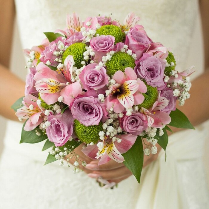 A radiant bridal bouquet, crafted with care near the leafy avenues of Golders Green, is gently clasped in a bride's hands, its round, abundant form set against the intricate lace fabric of an elegant wedding dress. The design centres on romantic roses, their petals in shades of soft pink and subtle lavender, some dewy and newly opened, others in full bloom-a celebration of tenderness and beauty. Green button chrysanthemums, their vibrant pom-pom shapes bringing a youthful spark, are placed throughout, introducing an unexpected brightness that recalls springtime strolls through Golders Hill Park. Pink alstroemeria, with striking dark pink and yellow markings, weave in an exotic charm, while billows of frothy white gypsophila fill the spaces with dreams of cloud-softness. Deeper green leaves anchor the delicate hues, lending fullness and structure to the bouquet. Every detail glows in the soft wedding light, from the subtle shine of a silver ring on the bride's finger to the careful flourish of each blossom. Designed by a Flowers Golders Green florist who understands the style and spirit of NW11, this bouquet encapsulates contemporary romance and timeless allure-an ideal choice for couples seeking fresh, inspiring wedding flowers for Golders Green and the surrounding area.