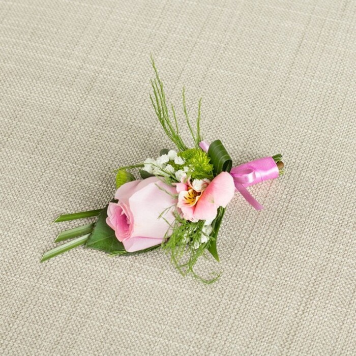 Beautifully set against the understated weave of light beige linen, this buttonhole arrangement radiates refined simplicity blended with artistic flair, an embodiment of Golders Green expertise. A central rosebud in muted baby pink, petals still tight and spiraling beautifully, rises with gentle poise, its core a subtle burst of deeper pink gradually diluting as it unfurls outward. Cradled to one side is a light peach-pink alstroemeria or lisianthus, petals opening playfully to reveal muted yellows and tiny maroon strokes, infusing the arrangement with intricate detail reminiscent of flower beds along Finchley Road. Accents of cloudlike baby's breath lace the bouquet, joined by vibrant green miniature cushion mums which enhance the boutonniere's sculptural feel. Layered beneath, feathery blades of dark grass, fern tips, and broad, well-folded leaves create visual structure and textural intrigue, suggesting both care and an artist's hand-hallmarks of local craftsmanship. At the base, lustrous satin ribbon in vivid fuchsia circles the stems and ties into a neat bow, lending a festive yet sophisticated finish. The combination of fresh, dew-kissed florals and lush foliage, illuminated by soft daylight and presented on understated neutral fabric, brings a sense of freshness and gentle celebration-ideally suited for weddings at Golders Green's historic venues, formal gatherings, or as a thoughtful accompaniment for local milestones.