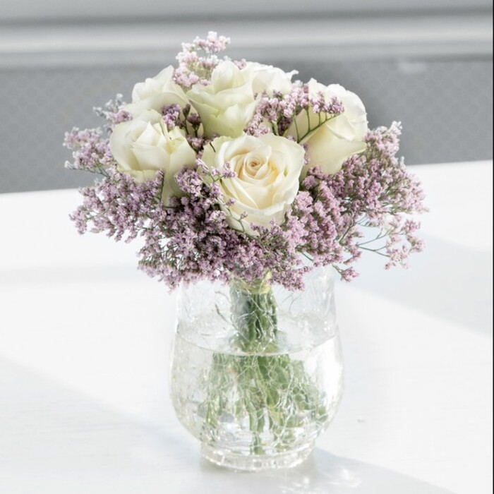 The photograph shows a beautifully balanced bouquet of creamy white roses and lavender statice, styled in a way that feels both contemporary and deeply romantic, much like the floral designs often chosen for homes around Golders Green and Hampstead. At the centre of the arrangement, around twelve white roses form a rounded crown. Their petals are velvety, with a soft ivory hue that catches the light in delicate gradients, from warm cream at the base to slightly lighter tones along the outer edges. None of the roses are fully blown; instead, each is at that perfect stage where the bloom is open enough to feel generous, yet still composed and elegant. Threaded throughout and surrounding the roses is a profusion of lilac statice, its tiny clustered flowers creating a misty, airy frame. The statice adds texture and a subtle lavender cast, like a gentle haze, softening the precise forms of the roses and lending the bouquet a dreamy, cloud-like quality. All the stems are gathered into a tall, clear cylindrical glass vase, where you can see the long, straight green stems submerged in calm, transparent water. The vase is placed on a spotless white surface that reflects the gentle daylight falling across the scene. The background is softly blurred, suggesting an interior with a large window, perhaps overlooking a quiet Golders Green garden or a leafy street near Brent Cross. The lighting is bright but diffused, giving the petals a luminous glow without harsh shadows. The mood is peaceful and refined, ideal for an elegant anniversary display, a thoughtful housewarming gift, or a calming sympathy bouquet that brings a sense of quiet comfort into any modern London home.