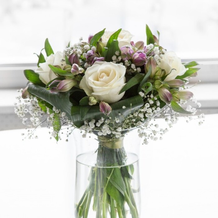 The photograph shows a beautifully balanced floral arrangement displayed in a tall, cylindrical clear glass vase, set against a bright, softly blurred background that feels like daylight filtering through a calm Golders Green living room. At the heart of the bouquet, classic white roses open in full, their petals layered in smooth spirals that create a plush, romantic texture. Each rose appears perfectly fresh, with edges untouched and centres softly glowing in the gentle light. Interlaced with these focal blooms are plentiful stems of lavender and muted purple alstroemeria, some still held in elongated buds, others beginning to unfurl into speckled trumpet-shaped flowers lined with delicate, darker veining. The purple tones offer a gentle contrast to the creamy white, bringing depth and a sense of quiet sophistication. Between these larger flowers, light, feathery sprigs of baby's breath scatter small white blossoms that resemble tiny clusters of stars, filling any gaps and adding an airy, cloud-like softness. Surrounding the blooms is a collar of rich green foliage, mixing broader, darker leaves with more slender, graceful stems that frame the arrangement and lend it a fresh-from-the-garden character, reminiscent of early morning walks near Golders Green Road. Inside the vase, the green stems stand straight and tidy in clear water, their different thicknesses and natural curves visible, highlighting the care and skill behind the composition. The lighting is calm and natural, enhancing the peaceful palette and evoking feelings of serenity, remembrance, and gentle joy. It is the sort of bouquet someone in Golders Green might have delivered to a loved one's home, equally suited for a thoughtful birthday, an elegant thank-you, or a tender sympathy gesture.