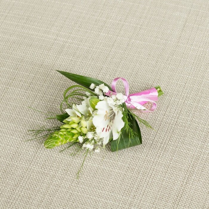 An artfully designed boutonniere is displayed on a softly textured, light beige fabric that resembles fine linen, the neutral backdrop giving the arrangement a serene, almost bridal quality. In the centre, a fully opened white alstroemeria commands attention, its petals slightly flared and velvety, with subtle curves that catch the light along the edges. Near the throat of the flower, fine reddish-brown speckles and streaks provide delicate contrast, drawing the eye inwards and highlighting the natural symmetry of the bloom. Just to one side, a compact, conical cluster of bright green buds forms a striking vertical accent, each bud tightly layered and overlapping like small scales, reminiscent of an Ornithogalum or ginger-style spike. This structural element brings a modern flair to the composition, playing against the softness of the alstroemeria. Woven throughout are tiny clusters of baby's breath, their miniature white flowers hovering lightly around the focal blossoms, softening any hard lines and adding a romantic, cloud-like veil. Supporting the flowers is a fan of broad, deep green leaves with pointed tips, providing a sturdy base, while fine, grass-like strands of greenery arc out from the sides and top, adding movement and a sense of organic flow, as though the piece has just been gathered by hand. At the right edge, a slim, glossy pink satin ribbon is tied into a precise bow, its vibrant hue and sheen injecting a joyful note into the gentle palette of whites and greens. The even lighting reveals every vein in the leaves and each speckled detail on the petals, emphasising the freshness of the stems. This boutonniere feels perfect for a groom or guest at a Golders Green ceremony, perhaps in a synagogue along Finchley Road or at a reception near Hampstead Heath, carrying a quiet promise of celebration and thoughtful local craftsmanship.