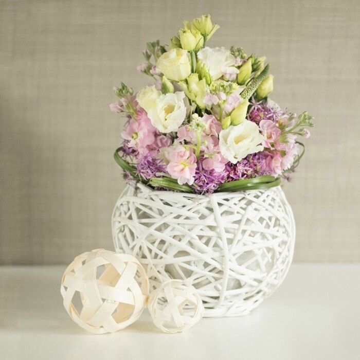 A beautifully curated still life, this abundant table arrangement captures the soft elegance that defines Golders Green's floral artistry. The spherical white vase-its open-work structure resembling a nest woven from painted rattan or slender branches-offers a light and contemporary frame. Overflowing with creamy white and pale yellow eustoma, each bloom showcases delicately layered, ruffled petals that evoke the gentle awakening of spring in NW11. Light pink and lavender-hued stock (matthiola) emerge above the main blossoms, adding graceful vertical lines and a soft, romantic fragrance. At the heart of the display, clusters of vivid purple filler flowers-likely petite asters or alliums-provide rich, velvety contrast and depth. Encircling the arrangement, a border of deep green foliage ties the colors together. Beside the vase, two woven decorative spheres in natural, bleached wood offer a subtle nod to Golders Green's mix of contemporary and earthy aesthetics. Set on a pristine white table and backed by a softly textured beige wall, the scene exudes tranquility and fresh sophistication. An ideal centrepiece for wedding celebrations, family gatherings, or Spring home refreshes, this composition radiates the expertise of local floral designers dedicated to merging tradition with modern flair in the heart of Golders Green.