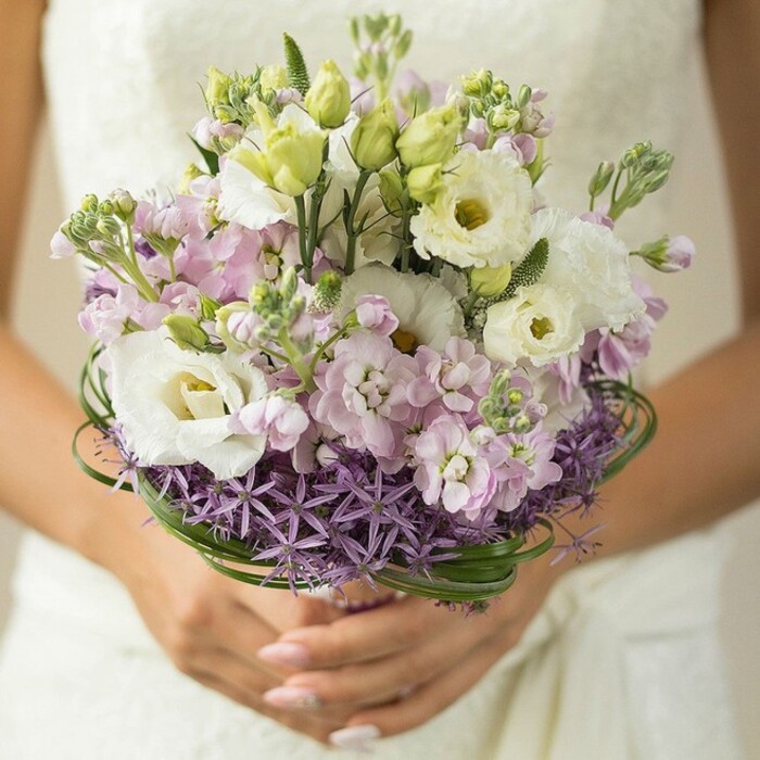 In this vivid close-up, a bride from Golders Green showcases her bouquet against the subtle backdrop of her lace ivory gown, the city's energy quietly present in the soft blur behind her. The arrangement embodies modern romance, designed with a harmonious balance of colours and textures popular throughout London's northwest neighbourhoods. Leading the composition are several large, snowy-white lisianthus flowers, their gently twisting, ruffled petals and luminous yellow-green cores drawing the eye. Around these, light pink and lilac stock blossoms cluster, offering depth, fragrance, and a sense of lush movement - the kind of tactile richness often found in local market bouquets along Finchley Road. Sprightly green buds and sinuous unopened lisianthus add freshness and a natural touch. At the base, a distinct band of deep purple, star-shaped allium delivers contrast and boldness, evoking the architecture and artistry unique to Golders Green's florists. Elegantly interwoven bear grass forms slim, glossy arcs just above the allium, defining and accentuating the bouquet's rounded shape. The bride's gracefully manicured hands, painted in a refined nude pink, steady the stems, while her dress's soft detail gently frames the composition. Every bloom is set to capture the joy of a spring or summer wedding in NW11, making this bouquet an ideal centrepiece for special occasions celebrated in the community.