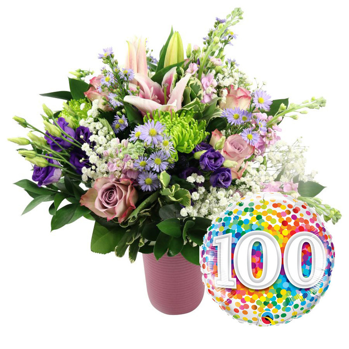 A striking centenary bouquet is showcased against a crisp white background, its colours and shapes captured with the clarity of a studio portrait. At the base, a cylindrical vase in muted dusty pink stands steady, its ribbed texture catching the light in gentle vertical lines that add a contemporary, tactile element. Rising from this vase, a lush arrangement of fresh flowers forms a rounded dome, overflowing slightly to create a sense of abundance. Several pink Oriental lilies command attention, some blossoms fully open, exposing their broad, star-shaped petals and prominent stamens, while others remain tightly furled buds that suggest ongoing life and growth. Around the lilies, an elegant mix of roses in light blush, mauve, and dusty rose shades spiral open, their soft, velvety petals giving the impression of something lovingly crafted by hand in a local Golders Green studio. Deep purple lisianthus blooms tuck between the roses, their silken petals introducing rich, jewel-toned depth. Fine sprays of dainty lavender asters, each with a small yellow heart, create a scatter of lighter texture, like tiny fireworks. Tucked among them are bright lime-green button chrysanthemums with tightly packed petals, lending a clean, modern accent. Wisps of white baby's breath cloud gently over the arrangement, mingling with glossy dark green foliage that frames the colours and provides structure. To the lower right edge of the bouquet, a large, round foil balloon leans into view, its metallic surface patterned all over with multi-coloured confetti dots in vivid reds, oranges, yellows, greens, blues, and purples. At its centre, bold white numbers spell out '100' with a faint shadow effect, making the milestone unmistakable. The entire composition speaks of a joyful 100th birthday or anniversary celebration, the kind of special moment families across Golders Green commemorate with flowers and heartfelt messages.