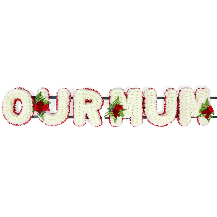 This dignified floral memorial spells out the words OUR MUM in bold, white letters that immediately catch the eye against a simple, uncluttered white background. Each letter is meticulously filled with small white chrysanthemum florets, tightly layered so that no greenery shows through, giving the surface a dense, cushioned look that appears soft to the touch. The petals form a uniform snowy texture across every letter, suggesting purity, peace, and a gentle farewell. Around the edge of each capital, a narrow but vivid border of deep red flowers defines and sharpens the outline. These red blooms, most likely spray carnations or miniature roses, run in an unbroken line along the perimeter of every letter, creating a pleasing contrast and enhancing the three-dimensional feel of the tribute. Behind the flowers, you can just make out the dark green supporting bars and frame that link the letters together, revealing the sturdy construction needed for display at a funeral service or burial, perhaps at a cemetery near Golders Green Road or Princes Parade. Adding depth, warmth, and a sense of personal affection, three clusters of red roses and fresh greenery are carefully woven into the letters. One rests between the O and U, another sits between the first M and the U, and the final grouping graces the far edge of the last M. The roses, with their velvety texture and rich colour, speak of deep and lasting love for a mother, while the green foliage and ferns introduce a natural, garden-like freshness. Overall, this OUR MUM floral tribute feels serene and heartfelt, a classic choice for families in Golders Green and nearby Hampstead Garden Suburb seeking a meaningful, beautifully crafted expression of remembrance.
