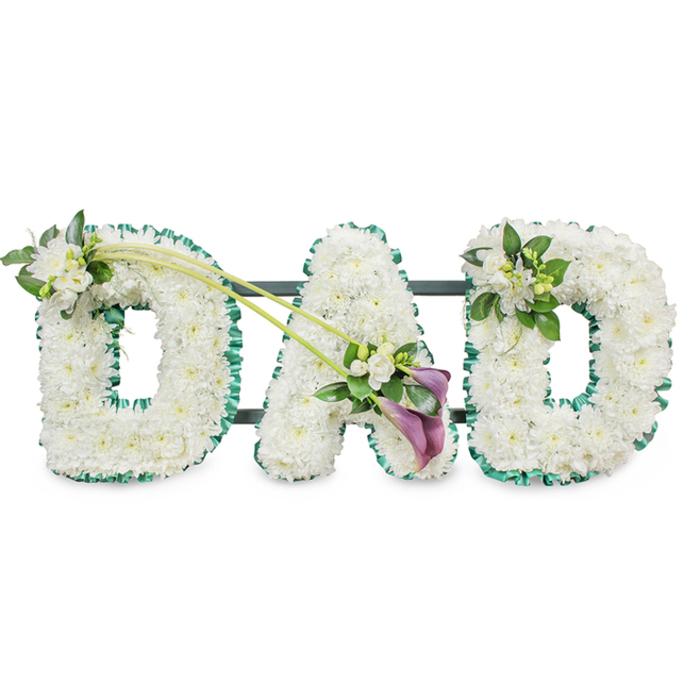 A solemn, meticulously assembled sympathy tribute forming the word DAD in large, three-dimensional letters-each letter composed of hundreds of small, immaculate white chrysanthemums or similar button blooms, tightly set to create a plush, velvet-like surface. A rich emerald-green satin ribbon is painstakingly applied around the outer edge of each letter, the folded ribbon providing crisp definition and a soft reflective sheen that contrasts with the matte texture of the flowers. The arrangement is supported by a dark frame that shows as horizontal bars behind the letters, confirming its upright display format for a chapel, funeral hall, or graveside. Floral embellishments are placed with sensitivity: the first D is graced at its upper left with a cluster of fragrant white freesia and lush green foliage; two elegant long stems curve from the top of that D toward the central A, finishing in an artful spray where two deep purple calla lilies stand amid more white freesia and leaves, their trumpet shapes adding solemn beauty and depth. The final D carries a slightly smaller grouping of freesia and greenery at its upper right, creating visual harmony. The quietly scented freesia and the soft feel of the chrysanthemums invite reflection. As Flowers Golders Green we prepare tributes like this daily for families attending services at Golders Green Crematorium, local churches, or community halls near Golders Hill Park, and we can arrange respectful delivery across Golders Green-see flowersgoldersgreen.mom for options and timings.