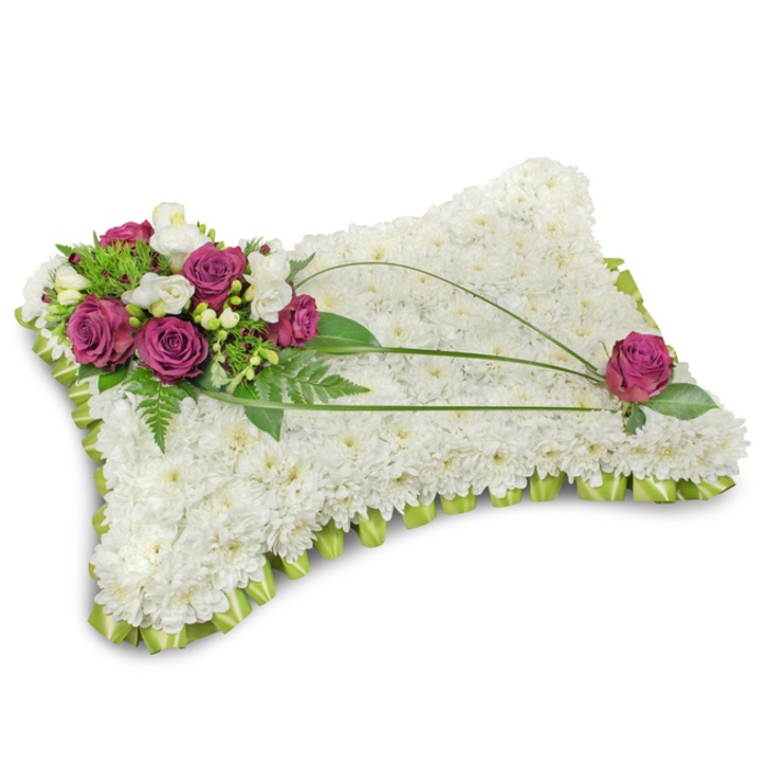 A traditional pillow-style condolence tribute, presented on a stark white background so every texture and hue reads clearly: the main body is densely covered in pristine white chrysanthemums, each tiny floret contributing to a plush, almost cloudlike surface that evokes peace and purity. Anchoring the top-left is an elegant cluster of deep magenta and fuchsia roses, their velvety petals unfolding beside delicate white freesia and soft, creamy lisianthus-which together lend a whisper of fragrance and a tender, refined contrast to the white field. Lush greenery, including fine fern fronds and broader glossy leaves, frames the cluster and adds dimensional depth while three slender bear grass stems arch gracefully from the focal grouping across the pillow, creating an understated motion that guides the eye. A single matching magenta rose is placed toward the lower-right corner, balancing the design and closing the visual loop. Around the base, a neatly folded lime-green satin ribbon forms a scalloped trim that both anchors the piece and adds a discreet flourish of colour. The studio light is gentle and diffuse, casting soft shadows that underline the cushion's contours. Crafted with sensitive attention to composition and symbolism, this floral pillow is an appropriate and dignified choice for services at Golders Green Crematorium, nearby synagogues, or intimate memorial gatherings close to Golders Hill Park.