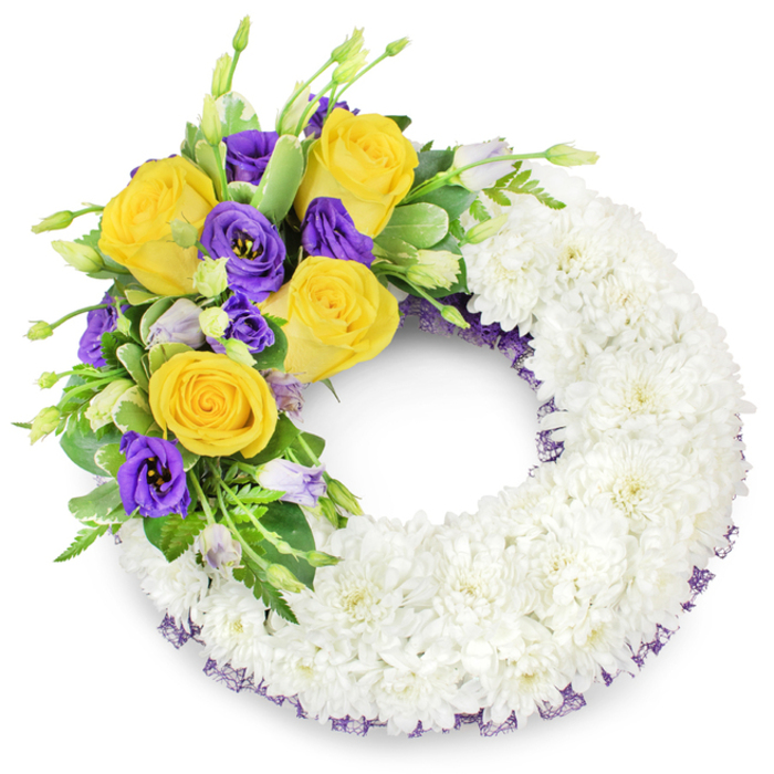 This image shows a traditional circular funeral wreath photographed against a plain white background, its craftsmanship evident in the compact, cloud-like ring of white spray chrysanthemums that forms the principal body. The chrysanthemums are densely clustered to create a smooth, reverent surface and a clearly defined open centre. From the upper-left portion of the ring a focused floral spray arcs gracefully toward the top, composed of several fully bloomed yellow roses with petals rolled into soft, golden spirals, accompanied by deep purple lisianthus (Eustoma) whose ruffled, satin-like petals introduce a sophisticated contrast. Interspersed among the primary blooms are a few lighter-lilac lisianthus buds and green buds that add dimensional layers and subtle colour transitions. Fresh greenery - including glossy, rounded leaves, delicate fern fronds, and slim stems of textured filler - frames the cluster and supports the blooms, helping them read clearly from a distance. A discrete trim of dark purple mesh ribbon is visible beneath the chrysanthemum layer along both inner and outer edges, a finishing touch that picks up the purple notes of the lisianthus and gives the piece a balanced silhouette. The lighting is soft and even, enhancing the chrysanthemums' matte texture and the roses' gentle sheen while keeping the overall effect calm and respectful. For families in Golders Green and surrounding neighbourhoods - often arranging tributes for services at Golders Green Crematorium or small memorial gatherings at home - this wreath reads as a composed, timeless expression of condolence and remembrance, handcrafted to convey dignity and lasting sympathy.