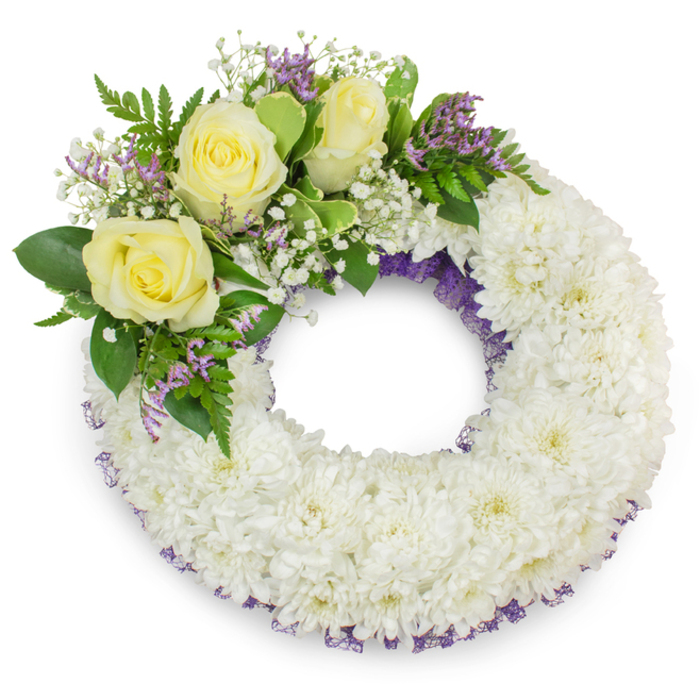 A composed, traditional sympathy wreath presented on a clean white field, its main structure made from densely packed white spray chrysanthemums whose many-layered petals form a plush, uniform ring. The wreath holds a measured, tactile presence - you can almost sense the cool, papery touch of each chrysanthemum petal and the muted, green-tinged aroma of fresh foliage. A whisper of pale purple netting or ribbon shows through the flowers along both inner and outer edges, adding an understated contour that enhances depth and solemnity. Placed on the upper-left quadrant, a cluster of three soft yellow roses opens with quiet grace; their velvety petals read as kindness and long friendship. Nestled among them, clouds of tiny white baby's breath (gypsophila) lend an airy, lace-like softness, while short sprays of lavender-purple statice or limonium give gentle points of contrast. The cluster is framed with mixed greenery - broad, glossy leaves and feathery fern-like stems - which bring a fresh, living green that stabilises the pale florals. Photographed in soft light, the wreath casts a delicate shadow that lifts it from the white backdrop and highlights the florist's careful placement and technical skill. As Flowers Golders Green would advise, this wreath is a classic choice for memorials and wakes in the local area - appropriate for chapel services near Golders Hill Park, the peaceful corners around Hoop Lane, or quiet graveside farewells by Finchley Road - and is prepared with sensitivity, ready for local delivery across Golders Green.
