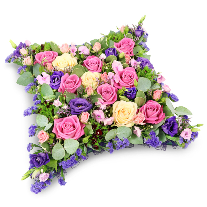 A carefully crafted floral pillow tribute, shaped like a soft rectangular cushion with gently rounded edges, lies in stillness against a seamless white background. The design is densely packed so that no greenery or stems are visible from above, just a rich tapestry of petals in pinks, creams, and purples. At the centre and radiating outward, large roses take pride of place: some are deep fuchsia, almost raspberry in tone, while others are tender blush pink or creamy pale yellow, their petals layered in spirals that suggest timeless romance and enduring affection. Scattered among these roses are deep violet and royal-purple lisianthus, their multi-layered, ruffled blooms adding drama and texture, like small evening clouds drifting across the arrangement. Between the main flowers, sprigs of tiny lavender-hued blossoms, resembling statice or limonium, infuse the tribute with a fine, almost feathery detailing that softens the overall look. Around and beneath the flowers, rounded eucalyptus leaves in a muted silver-green tone and darker, slender fern-like foliage peek through, offering contrast and a cool, fresh undercurrent, as if echoing the tranquil green spaces around Golders Hill Park and the quiet residential streets of Golders Green. The crisp white background throws the colours into sharper relief, emphasizing the subtle height and cushion-like dome of the design, while a delicate shadow suggests gentle, diffused lighting. This floral pillow feels deeply contemplative, the sort of tribute a family in Golders Green might select for a funeral, cremation, or graveside service when they wish their final message to be one of serene rest, gratitude, and loving remembrance.
