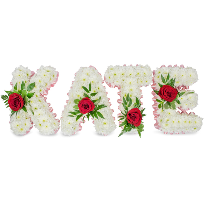 This image presents a considered and elegant floral name tribute spelling KATE in large, three-dimensional block letters, each letter painstakingly formed from a voluminous cluster of small, pristine white chrysanthemums. The chrysanthemums create a soft, pillowy texture that suggests purity and dignity, and along the outer edges each letter is finished with a subtle pale pink satin ribbon that is pleated into a neat, delicate border. A single, vivid red rose sits prominently on each letter as a bold point of contrast: on the K the rose is arranged on the lower part of its left vertical stroke; on the A the rose is positioned at the apex of the triangular peak; on the T the rose rests at the base of the left section of its horizontal crossbar; and on the E the rose graces the middle horizontal bar. Around each rose, lush green foliage - a combination of broad glossy leaves and finer fern-like sprigs - adds depth, framing the crimson blooms and softening the hard geometry of the letters.

The clean white backdrop and gentle, diffuse lighting allow the textures and colours to speak clearly: satin whisper ribbons, the chrysanthemum's layered petals, the velvety crimson of the roses and the cool sheen of the greenery. The arrangement reads as a bespoke sympathy tribute suited to funerals, memorials or remembrance ceremonies where clarity and heartfelt sentiment are required. As a local piece one might commission from a Golders Green florist working near Golders Hill Park or Hendon Lane, it balances traditional floral symbolism - white for purity, red for love - with meticulous florist craftsmanship to honour a life with quiet, assured grace.