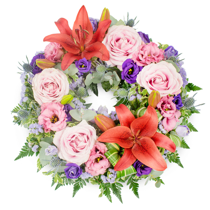 This image shows a beautifully crafted funeral wreath laid flat against a clean white background, its circular form symbolising eternity and the enduring nature of love and memory. The wreath itself is generously filled, with every part of the ring covered by flowers and foliage so that it appears lush and complete. Soft blush-pink and light lavender-pink roses are placed throughout, many of them fully open so that their delicate, layered petals form gentle spirals. These roses give the arrangement a romantic softness and a sense of calm remembrance. Interspersed among them are eustoma, or lisianthus, in two main tones: ruffled blossoms of pale pink and more dramatic, deep purple blooms that add depth and visual contrast. Two prominent Asiatic lilies in a vivid coral-orange shade provide strong focal points, one positioned at the upper portion of the wreath and the other near the lower right. Their broad, star-shaped petals and clearly defined stamens bring an energetic, almost sunlit warmth to the design, offsetting the cooler pinks and purples. Around them, several unopened lily buds peek from the foliage, suggesting new life and continuity. The floral ring is supported by a rich bed of greenery: rounded, silvery-green eucalyptus leaves create a soft, muted backdrop; dark, slender fern fronds introduce movement and light feathery lines; and other textured leaves complete the frame. Distinctive blue-grey eryngium thistles with spiky heads punctuate the foliage, lending an architectural, modern touch. Threaded throughout are tiny lavender-blue filler flowers, likely statice or limonium, which create a fine, misty veil of colour. Small clusters of bright green, carefully folded leaves near the bottom showcase expert floristry techniques. The overall feel is peaceful yet expressive, a thoughtful wreath that could easily be imagined resting at a service in one of Golders Green's local synagogues or chapels, offering a final message of respect and sympathy.