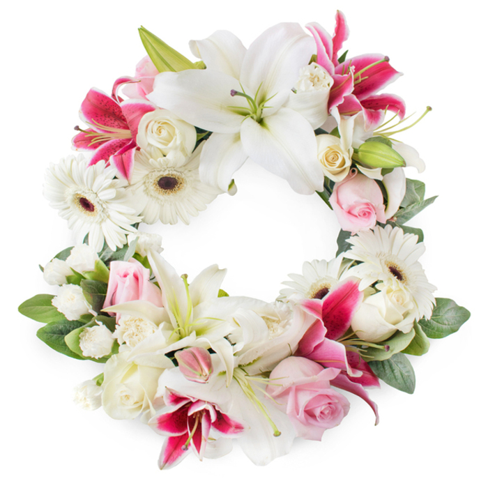 Set against a bright white background, this open circular wreath appears as a serene floral ring, richly packed with white, soft pink, and deep magenta blooms, designed as a tender farewell piece. The most prominent feature is the cluster of large white lilies spaced around the circle, their generous petals opening outward in a graceful star, revealing pale green centres and fine golden stamens that give the arrangement a refined, almost luminous quality. Among them, deep pink and fuchsia lilies weave in and out, their richer tones and intricate speckling adding depth and a note of passionate remembrance that contrasts beautifully with the quieter whites. Nestled closely between the lilies are groups of soft pink roses, some still cupped in tight buds, others partially unfurled, suggesting gentleness and affection. Creamy white roses, fully open with densely layered petals, add volume and a classic, timeless feel often chosen for funeral tributes in the Golders Green area. White gerbera daisies, with their layered petals and distinctive dark brown centres, bring a slightly modern, uplifting touch that softens the formality of the lilies and roses. Smaller white filler flowers, such as miniature carnations or spray chrysanthemums, tuck into the spaces, creating a cushion of delicate texture that makes the wreath appear full and lovingly composed. A backdrop of varied green leaves curves around the blooms, framing the circular shape and adding fresh, garden-like depth. This thoughtfully arranged wreath would be fitting for a service at a local chapel near Golders Green station or a memorial gathering at Golders Hill Park, symbolising eternal love, peace, and enduring memory.