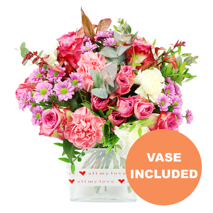 A vibrant mixed flower arrangement takes centre stage, carefully styled in a clear, rectangular glass vase and photographed against a bright white backdrop so every hue is crisp and true. This Golders Green bouquet is dominated by generous pink roses, some in intense fuchsia with almost velvety, darker outer petals, others in gentle blush or pastel tones, their spiralling layers opening like small, romantic sculptures. Surrounding and supporting them are plush carnations in soft coral, peach-pink, and creamy white, their characteristic ruffled petals adding fullness and a tactile richness that almost invites you to reach out and touch. Between the larger blooms, petite daisy-like chrysanthemums in lavender and light purple punctuate the design, their neon-green centres acting like tiny sparks of energy that enliven the overall palette. The flowers are anchored by a tapestry of foliage: rounded eucalyptus leaves with a cool, silvery-green cast, darker glossy greens that give depth, and a few hints of reddish-brown foliage that lend a gentle, autumn-tinged note, reminiscent of an early evening walk through nearby Golders Hill Park. The transparent vase, with its clean, modern silhouette and softly rounded corners, is wrapped near the base with a white band decorated in repeating bright red hearts and the romantic phrase all my love in flowing red script, visually declaring this as a message of affection. In the lower right corner of the image, an orange circular label stands out, clearly printed with VASE INCLUDED in bold white capital letters to highlight the convenience of receiving a complete, ready-to-display gift. The overall effect is joyful and heartfelt, an ideal arrangement for anniversaries, Valentine's Day, Mother's Day, or simply sending love with same-day delivery along Finchley Road and into the surrounding streets.