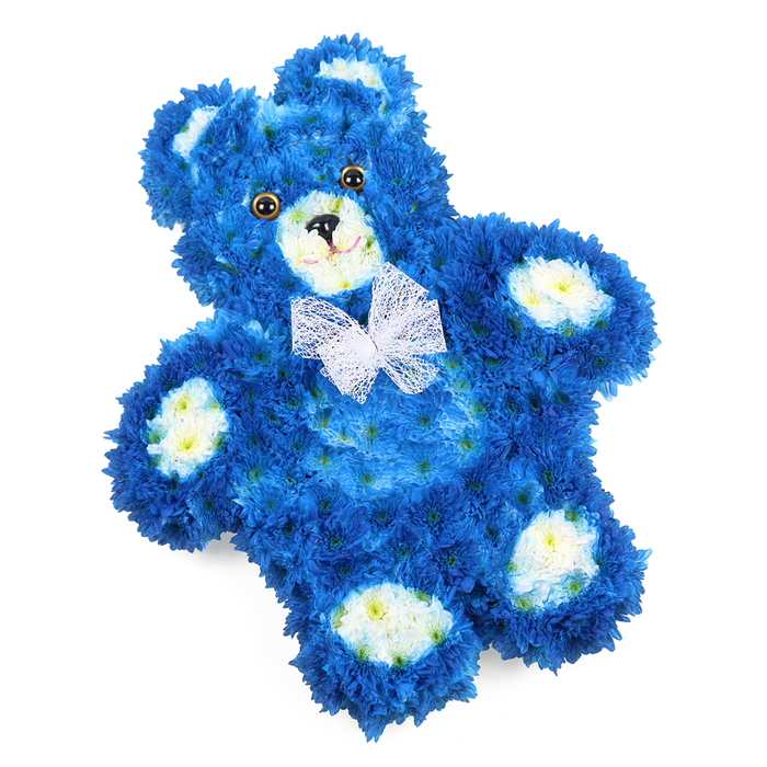In this image, a floral teddy bear lies flat yet appears wonderfully three-dimensional, its entire body composed of densely packed flowers arranged with the precision of an experienced florist. The main colour is a sumptuous royal blue, created by rows of tiny chrysanthemum-like blossoms nestled tightly together so no gaps in the structure are visible. This deep blue coating wraps seamlessly over the round head, plump belly, and outstretched limbs, giving the impression of a soft, plush toy ready to be hugged. Cream and soft buttery-yellow blooms are carefully placed to form the rounded muzzle, the inner curves of both ears, and the four oval pads on the paws, creating gentle contrast and highlighting the classic teddy silhouette. The face is lovingly detailed: two large, polished-looking eyes in a dark golden-brown appear slightly reflective, adding life and personality, while the small black triangular nose and the fine curved line beneath it form a subtle, peaceful smile. Around the bear's neck, a delicate white mesh bow sits just so, its airy loops and soft tails resting lightly against the blue petals, as if tied with quiet ceremony before a service. Set against a pure white backdrop, every petal and contour stands out, showcasing the craftsmanship that might be expected from a specialist florist serving the Golders Green area and nearby Hampstead Garden Suburb. This floral teddy bear feels particularly fitting as a tribute for a baby boy or young child, or as a gentle sympathy arrangement sent to a family home or chapel, offering a symbol of innocence, comfort, and lasting love expressed through fresh seasonal flowers.