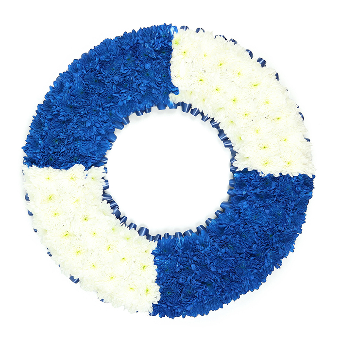 This image shows a lifebuoy-inspired funeral wreath, perfectly round and presented against a spotless white backdrop so that every detail of the flowers stands out clearly. The ring is thick and substantial, built from hundreds of small, uniform blossoms that have been placed very close together, leaving no gaps and producing a soft yet structured texture. The colour layout follows a distinct, quartered pattern: starting at the upper left, a block of rich royal blue blooms leads into a segment of bright white flowers at the top right, then back to royal blue at the lower right, and finally to white again at the lower left. Each quadrant stops at a crisp, straight boundary, giving the arrangement a bold, graphic look that still feels gentle thanks to the rounded shapes of the petals. Inside the central opening, a narrow, dark blue ribbon runs around the inner circumference, gathered into fine ruffles that gently echo waves lapping around a buoy at sea. There is no foliage or additional adornment, only these blue and white flowers, chosen for their resilience and ability to stay fresh and vibrant during a service or graveside farewell. The overall mood is respectful and quietly maritime, ideal for commemorating someone in Golders Green with ties to the navy, merchant shipping, or simply a deep love of the coast and open water. The tribute could easily be imagined travelling from a local shop near Golders Green Station to a family home or service venue, offering a strong visual message of safety, remembrance, and enduring love. Every curve and colour block reflects the steady hands and thoughtful eye of a professional florist, creating a meaningful symbol of hope and final rest.