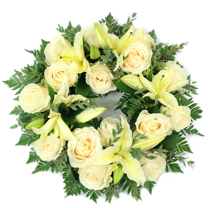 A dignified circular wreath, carefully photographed from above, composed of creamy, near-ivory roses and soft pale yellow lilies set into a rich, verdant cushion of foliage. The design is dense and balanced: around fifteen or so fully opened roses with neatly coiled petals form the primary visual rhythm, their soft cream catching light to create a serene, almost luminous band. Scattered among them are elegant trumpet lilies in a muted lemon shade; some are fully unfurled, their elongated petals and subtle yellow stamens exposed, while others remain as supple buds, introducing vertical nuance and the sense of unfolding life. The greenery is deliberate and textural - light, feathery fern fronds lend a softened perimeter, while broader, rounded leaves, likely eucalyptus, provide cool bluish-green tones and structural contrast. The overall colour story of ivory, pale yellow, and layered greens reads as composed and consoling. Sensory details come through in the description: the roses' gentle, powdery sweetness and the lilies' fresher, green-tinged fragrance would mingle softly without overpowering the room. Presented flat against a white background, the continuous circular form symbolizes eternity and remembrance, making the wreath especially fitting for a funeral service, memorial tribute, or grave-side offering. As a local piece crafted for families around Golders Green and nearby Hampstead Heath, it embodies restraint, respect, and the quiet comfort of an experienced florist's hand.