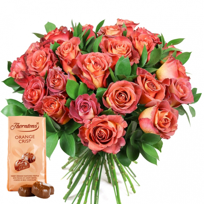 A showstopping bouquet and gourmet chocolate set, gracefully arranged by dedicated florists from Golders Green, takes centre stage in this luminous image. The eye is immediately drawn to the lavish sweep of fresh roses, clustered closely together and alive with tones of coral, deep rose, golden peach, and subtle pinks. Many blossoms showcase a gorgeous bicolour gradient-centres that glow with creamy yellow and soft peach, with saturated, rosy-pink tips accentuated on the fringes. The intricate, velvety texture of each petal is captured in perfect detail, inviting both admiration and the imagined softness of a freshly cut rose. Nestled among the flowers, rich green foliage-likely ruscus or eucalyptus-offers refreshing contrast and grounds the arrangement in nature, echoing the leafy avenues near Golders Green station. At the front, complementing this floral masterpiece, stands a light cream Thorntons Orange Crisp chocolate pouch, upright and clearly branded. Its surface is adorned with images of glossy chocolate bites and text describing the truffle's tantalising orange-flavoured filling and crispy centre, conjuring a sensory anticipation. Two actual truffles rest gracefully in front: spherical, dark, and gleaming. This refined gift set, with its interplay of warm florals and indulgent chocolate, suggests thoughtful gifting for occasions like Mother's Day, anniversaries, or celebrations near Brent Cross, all beautifully presented for delivery throughout Golders Green's distinctive neighbourhoods.