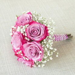 This radiant bouquet in Golders Green captures the essence of artisan floristry, inviting you to savour its nuanced textures and carefully harmonised palette. Central to the arrangement are radiant roses, their petals displaying a captivating gradient: velvety lavender and mauve interiors deepening to bold magenta at the outer edges. Delicate baby's breath weaves a cloudlike counterpoint through the clusters, its white florets softening the composition with a touch of ethereal beauty. The bouquet is gracefully bound in a stream of deep purple satin ribbon, adorned with spiraling, iridescent pearl strands-a luxurious detail reminiscent of classic Finchley Road boutiques. The stems, freshly cut and visible beneath the ribbon, affirm the bouquet's vitality. Sunlight from a nearby window casts a gentle glow over the soft beige, linen-textured surface below, making every colour and texture stand out. There's an aura of joyful expectation and refined celebration, perfectly suited to Golders Green weddings or intimate gatherings. This bouquet weaves together the local spirit, contemporary design, and traditional floral elegance, inviting you to imagine its gentle fragrance enhancing a joyous day in this leafy London suburb.