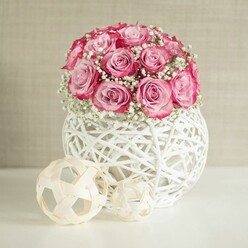A refined floral centerpiece sits poised on a clean white surface, as if styled for a Golders Green townhouse just off Finchley Road, showcasing a rounded bouquet of pink roses cradled by clouds of white baby's breath. The roses are tightly clustered, each bloom displaying a beautiful gradient: petals blush softly at the center before shifting to a richer, deeper pink at the edges, with hints of fuchsia where the light catches the curves. Between the roses, sprays of gypsophila thread through like airy lace, their countless tiny white blossoms adding lightness and a sense of quiet celebration. The bouquet emerges from a distinctive spherical vase, crafted from white-painted, woven strands that mimic wicker or rattan, its open lattice design creating a sense of air and transparency. Light filters through the criss-cross pattern, casting subtle shadows on the smooth surface below and reinforcing the airy mood. In front and to the left of the vase rest two decorative balls in soft natural tones, made from slim interlaced wooden strips; their geometric, open shapes create a visual echo of the vase's texture and introduce a touch of minimalist, Scandinavian-style décor. The background is a softly textured wall in warm beige or cream, unobtrusive yet comforting, like the quiet interior of a well-loved local café on Golders Green Road. Gentle, diffused lighting bathes the arrangement, enhancing the pinks and whites without harsh contrast, so the overall effect is soothing, romantic, and contemporary. This scene evokes a thoughtful gift for a birthday, new baby, or intimate dinner at home, crafted with the care you would expect from an experienced local florist.