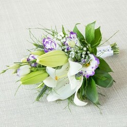 An elegant, elongated bouquet lies at a gentle diagonal on a pale beige, softly woven fabric, like a bridal arrangement placed on a dressing table before a ceremony in Golders Green. At the heart of the design are several large Oriental lilies, their white petals smooth and almost velvety, opening wide to reveal strong yellow-orange stamens and subtle freckles of warm reddish-brown deep inside the throat of each flower. Nearby, smooth, tapered lily buds in bright, leaf-green tones stretch outwards, still closed and plump, lending a sense of anticipation and future bloom. Between the lilies, Eustoma (lisianthus) blossoms unfurl, their petals delicately crimped and ruffled at the edges, coloured in creamy white that melts into vivid purple margins and deeper violet centres, giving the arrangement a contemporary twist against the classic white. Clouds of tiny white baby's breath (Gypsophila) are scattered throughout, their minute blossoms like stars against the richer flowers, softening the lines and bringing a light, airy romance. The greenery is abundant yet carefully placed, with broad, dark green leaves forming a strong base and slimmer, more feathery foliage and spiky accents splaying outwards, creating texture, rhythm, and a sense of natural movement. At the tapered end, the exposed stems are bound tightly in a smooth white satin ribbon, finished with a sprinkling of small pearl-like beads that catch the light in a subtle, understated way, hinting at weddings, anniversaries, or milestone celebrations. The soft, even lighting and neutral backdrop allow every petal, leaf, and bead to be clearly defined, making this bouquet ideal for describing a classic yet modern wedding arrangement available for same-day delivery around Golders Green, Temple Fortune, and nearby Finchley Road venues.